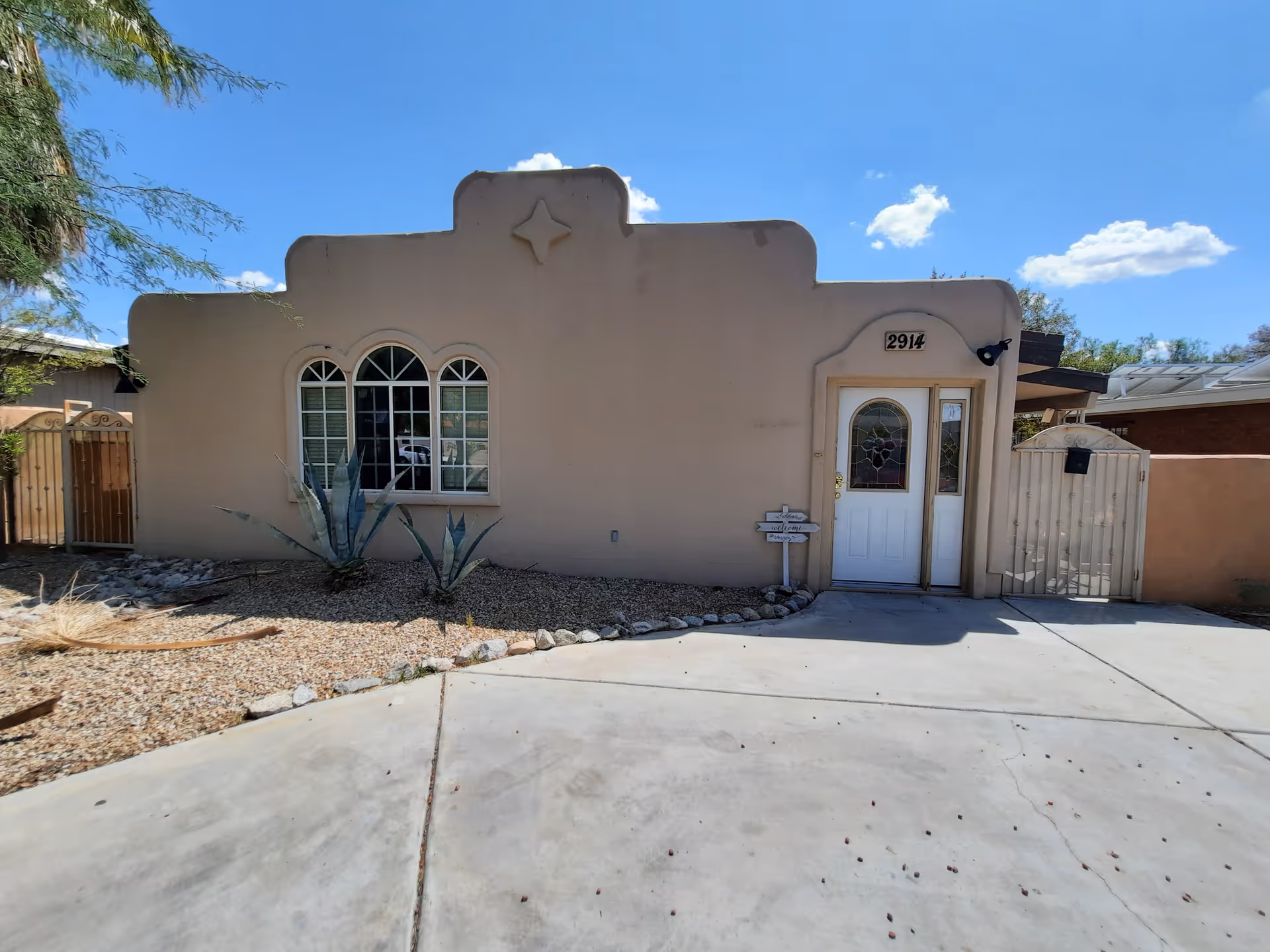 Exterior front view of a single-story building with a beige stucco facade, arched windows with white frames, a white door with decorative glass panels, and a small gated entrance on the right. The building number 2914 is displayed above the door. The ground is covered with gravel and desert plants, and the sky is clear with a few clouds.