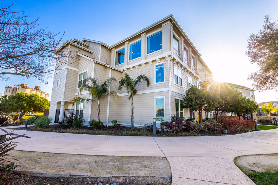 Exterior view of a multi-story beige senior living facility building with large windows, surrounded by palm trees, shrubs, and a paved walkway under a clear blue sky with the sun shining brightly.