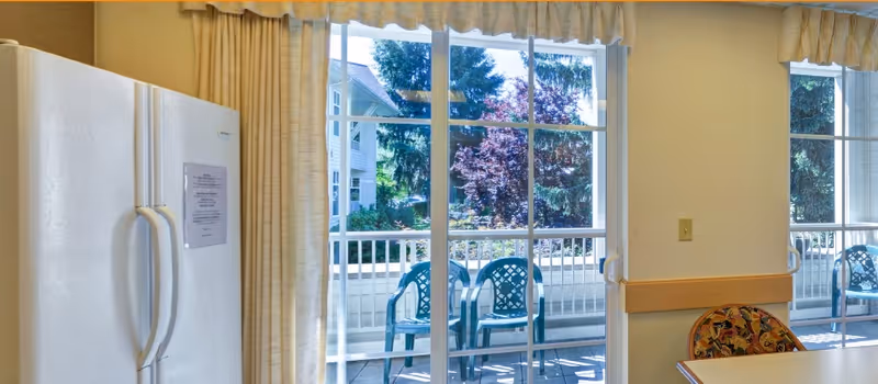 Interior dining area with a refrigerator and a patterned chair beside large sliding glass doors opening onto a patio with plastic chairs.