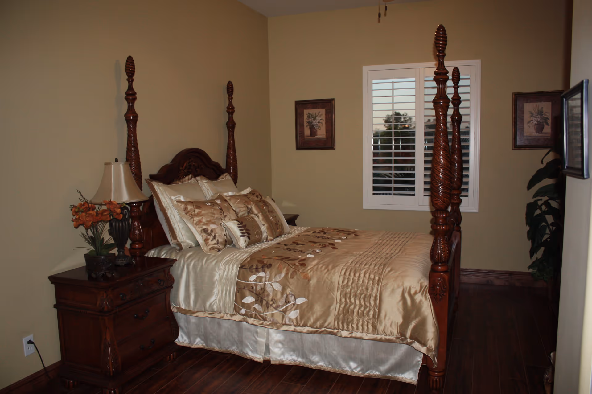 A bedroom with a wooden four-poster bed adorned with beige and brown floral bedding. There is a wooden nightstand with a lamp and a small flower arrangement on the left side of the bed. The room has beige walls, a window with white shutters, two framed floral pictures on the walls, and wooden flooring.