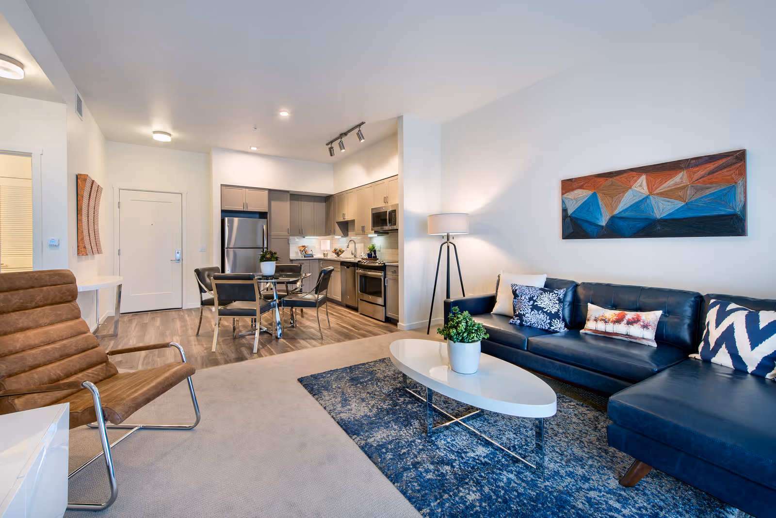 Modern open-concept living area with a dark blue sectional sofa adorned with patterned pillows, a white oval coffee table with a potted plant, a floor lamp, and a colorful geometric wall art. Adjacent to the living area is a kitchen with stainless steel appliances, gray cabinets, a small dining table with four chairs, and wood flooring.