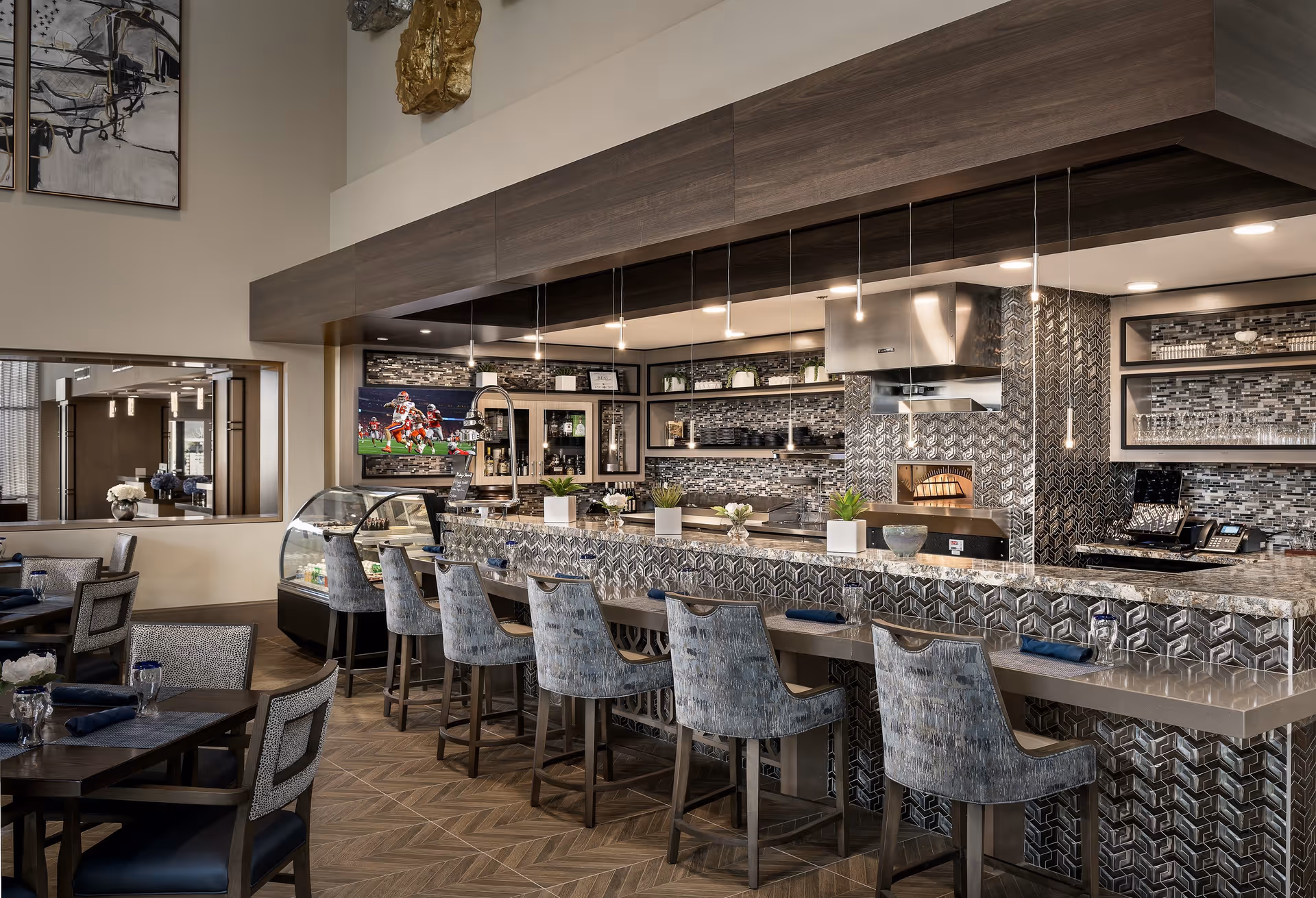 Modern dining area with a long counter featuring six upholstered bar stools, a granite countertop, and a decorative tiled backsplash. Behind the counter is a kitchen area with a pizza oven, shelves with plants and dishes, and a mounted TV showing a football game. Several dining tables with chairs and place settings are visible in the foreground.