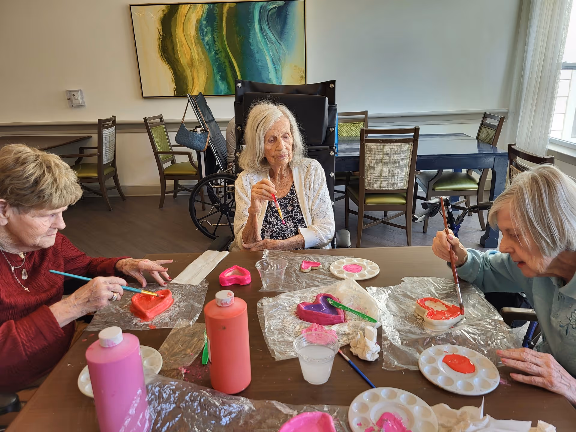Three elderly women sitting around a table in a well-lit room, painting heart-shaped objects with pink and red paint. Various painting supplies, including paint bottles, brushes, and palettes, are spread out on the table. One woman is in a wheelchair, and there are chairs and tables in the background along with a large abstract painting on the wall.