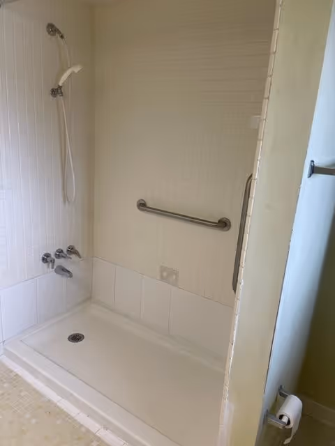 A shower area with white tiled walls and a white shower base. There is a handheld showerhead attached to the wall, two faucet handles, and a grab bar mounted horizontally on the wall for support. A toilet paper holder is visible on the right side near the entrance.