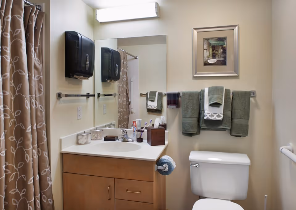 A clean bathroom with a white toilet and a wooden vanity with a white countertop. On the countertop are a toothbrush holder with toothbrushes, a tissue box, and containers. Above the vanity is a large mirror with a wall-mounted light fixture. To the left, there is a shower curtain with a leaf pattern. On the wall above the toilet, there are three green towels with a white decorative towel in the middle, and a framed picture above them. A black wall-mounted soap or paper dispenser is visible next to the mirror.