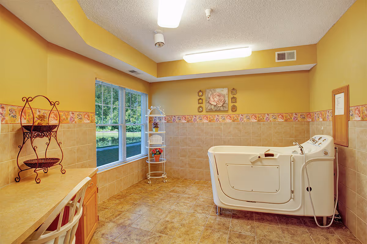 A spacious bathroom with yellow walls and tiled lower walls and floor. There is a large walk-in bathtub with a door on the right side, a window with blinds on the left wall, a small white shelving unit with potted plants, and a countertop with a chair and decorative metal stand. The room is well-lit with ceiling lights.