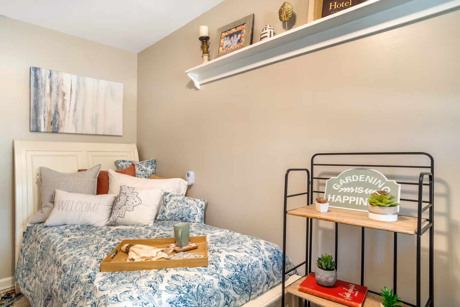 A cozy bedroom with a bed featuring a blue and white patterned comforter and multiple pillows, including one with the word 'WELCOME'. A wooden tray with a cup and napkin is placed on the bed. On the right side, there is a black metal and wood shelf holding small potted plants and a sign that reads 'GARDENING IS HAPPINESS'. Above the bed, a white shelf displays decorative items and a framed photo. The walls are painted beige and there is a piece of abstract artwork hanging above the bed.