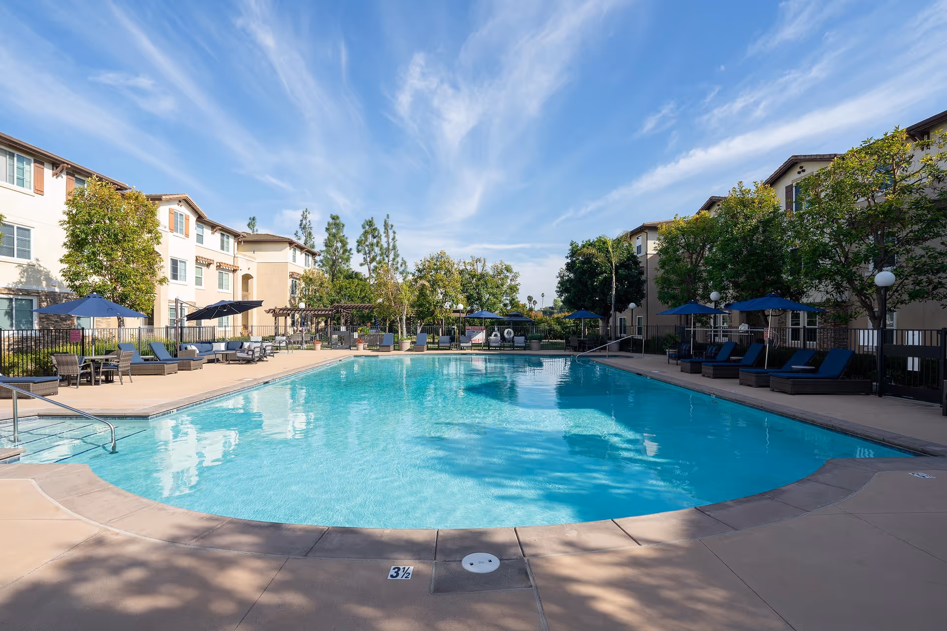 Outdoor swimming pool surrounded by lounge chairs with blue umbrellas, trees, and multi-story residential buildings under a partly cloudy blue sky.