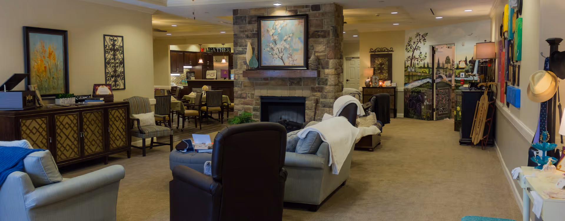 A cozy senior living facility common area with comfortable seating including armchairs and sofas arranged around a stone fireplace. The room features warm lighting, decorative paintings on the walls, and a dining area with tables and chairs visible in the background. The space is decorated with various homey touches such as blankets, cushions, and wall art, creating a welcoming atmosphere.