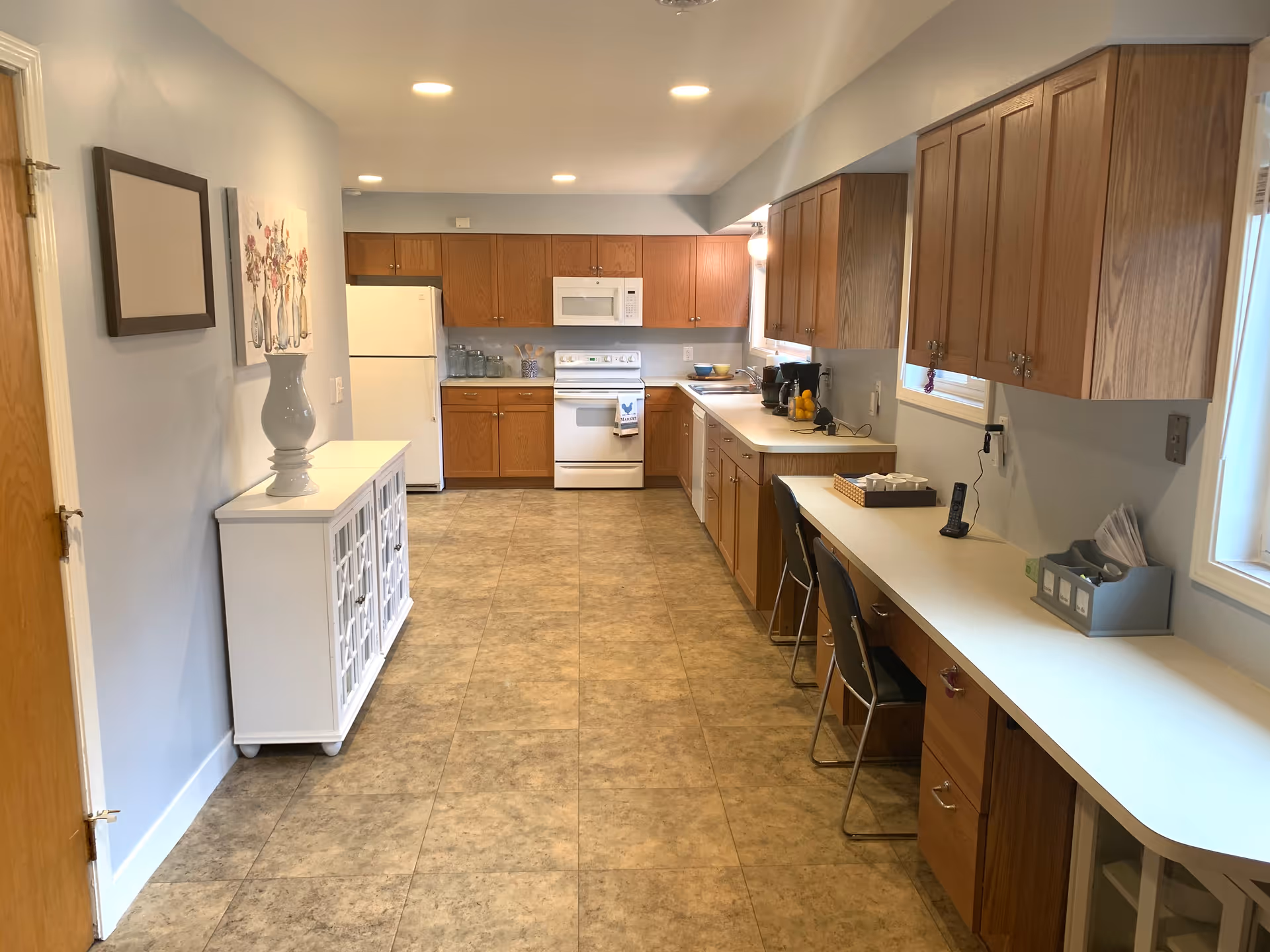 A spacious kitchen with wooden cabinets, white countertops, and tiled flooring. The kitchen features a white refrigerator, stove, and microwave. There are two chairs placed at a long counter on the right side, which also holds a phone, coffee maker, and other small items. On the left side, there is a white cabinet with a large vase and a floral painting above it. The room is well-lit with recessed ceiling lights and natural light from windows.