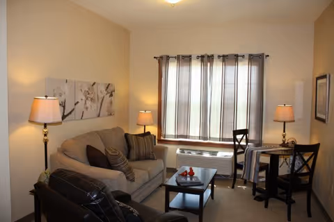 A cozy living room with a beige sofa adorned with multiple cushions, a dark leather armchair, a glass coffee table with decorative items, two floor lamps providing warm lighting, a small dining table with two chairs near a window with sheer curtains, and neutral-colored walls with artwork.