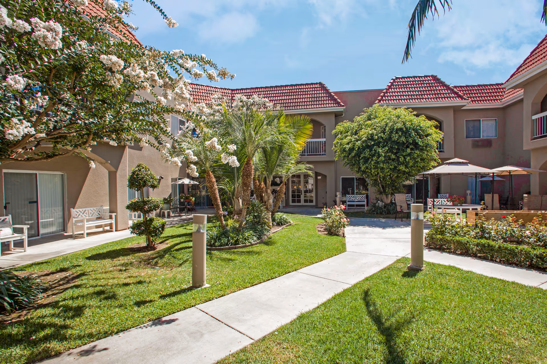 Sunny landscaped courtyard with walkways, palm trees, benches, and two-story buildings with red tile roofs.