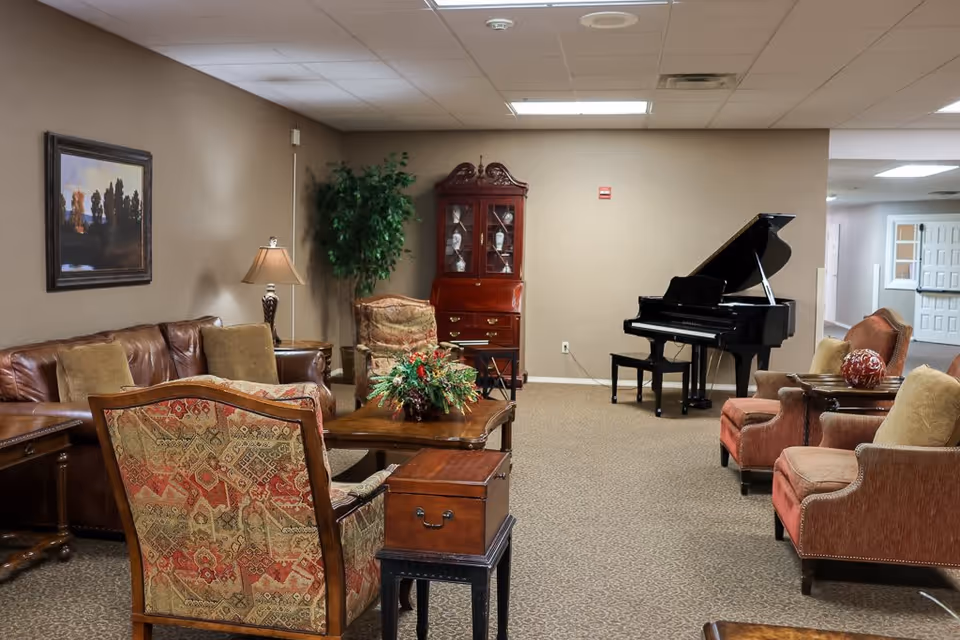 A cozy assisted living facility common area with a brown leather sofa, patterned armchairs, a wooden coffee table with a floral arrangement, a floor lamp, a potted plant, a wooden cabinet, and a black grand piano against a beige wall.