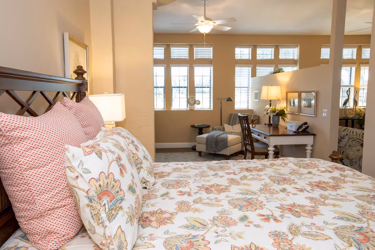A cozy bedroom with a bed featuring floral and patterned pillows and bedding. The room has beige walls, a wooden headboard, a bedside lamp, a desk with a chair, a table lamp, and a comfortable armchair near large windows with blinds allowing natural light to fill the space.