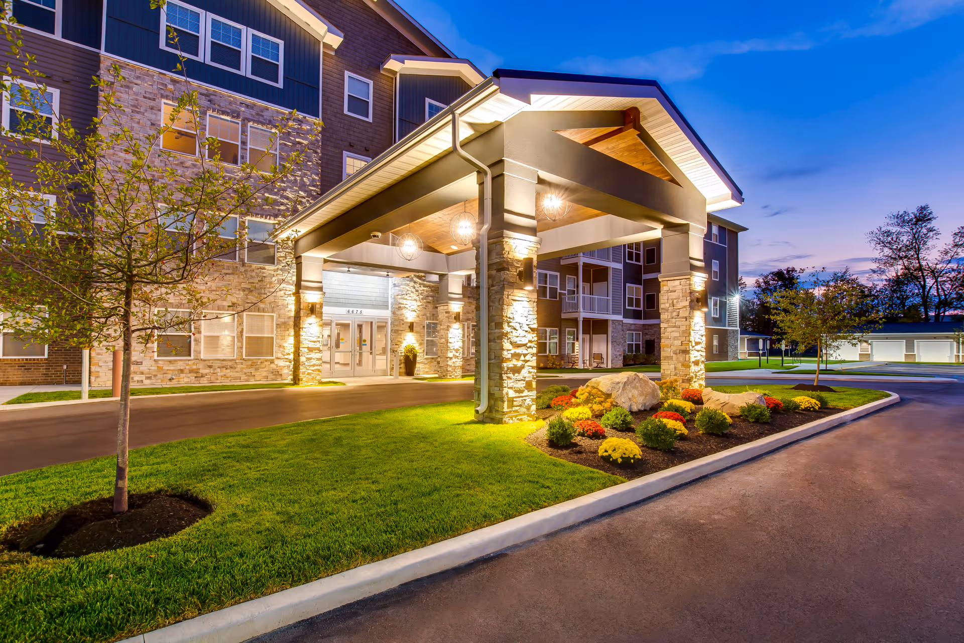 Exterior view of Encore 55+ Boutique Apartments in Plainfield at dusk, showing a well-lit entrance with stone pillars, a covered driveway, landscaped greenery, and a multi-story building in the background under a clear blue sky.