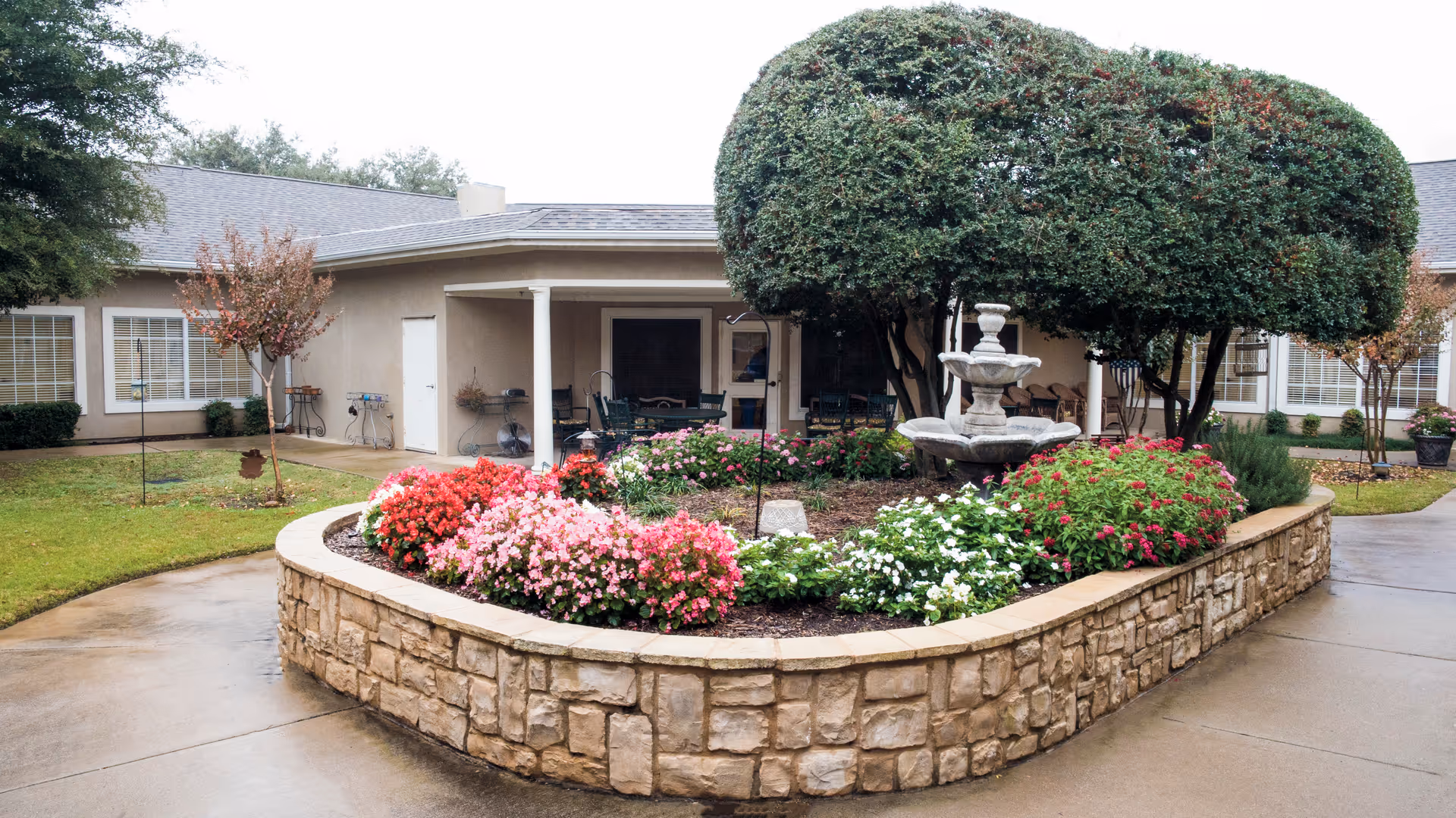 Stone raised flowerbed filled with colorful flowers and a tiered fountain in front of a single-story building with a covered patio and trimmed trees.