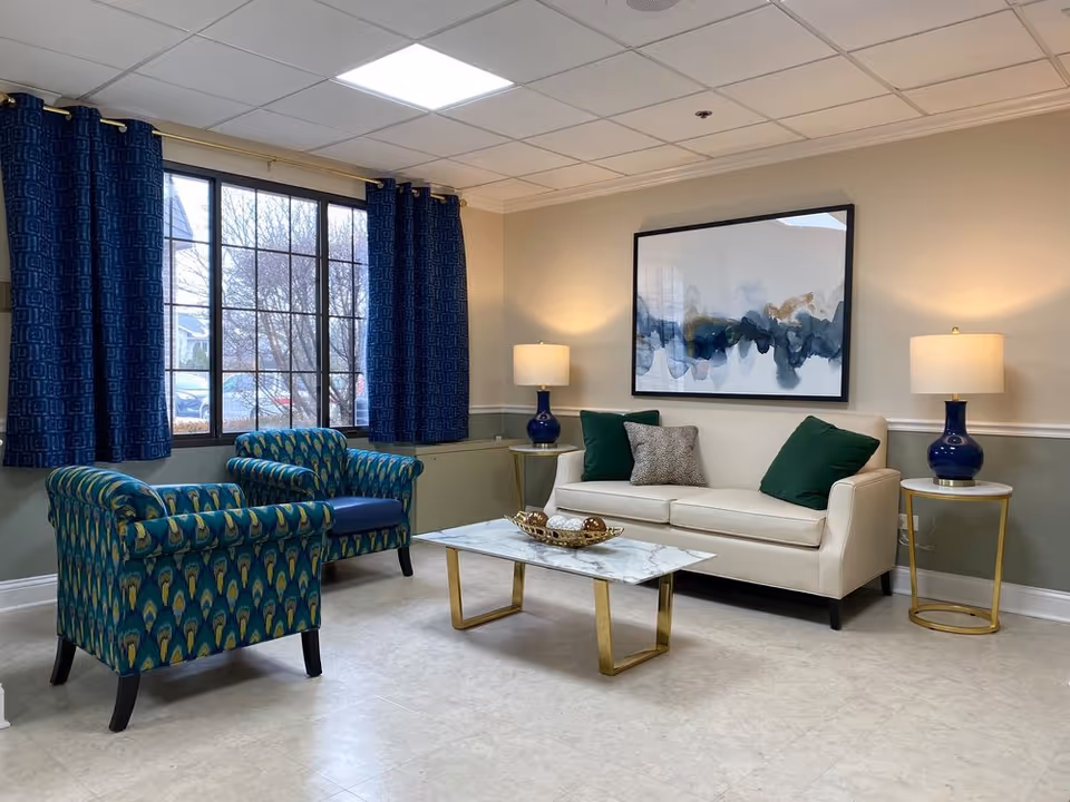 A cozy living room with a beige sofa adorned with green and patterned pillows, two blue patterned armchairs, a marble coffee table with decorative items, two side tables each with a blue lamp, and a large window with dark blue curtains letting in natural light. A large abstract painting hangs on the wall above the sofa.