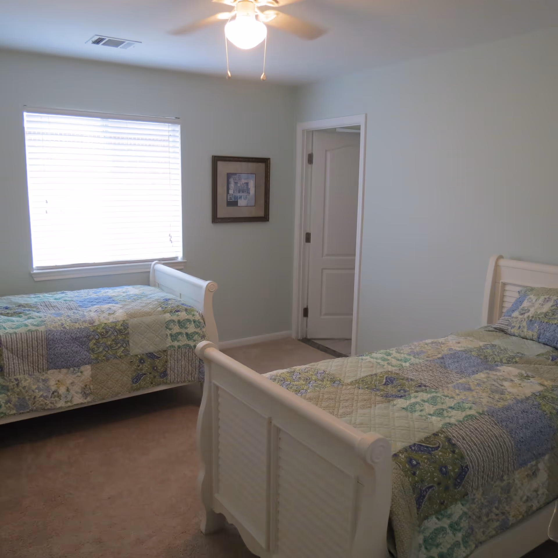 A bedroom with two white wooden twin beds, each covered with a patchwork quilt in shades of blue, green, and beige. There is a window with closed blinds on the left wall and a framed picture hanging on the wall between the window and an open door. The room has light-colored walls and carpeted floor, with a ceiling fan and light fixture above.