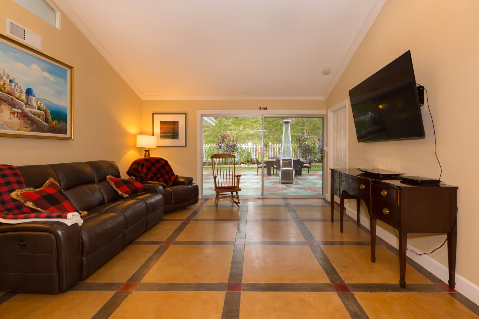 A cozy living room with a brown leather sofa and armchair adorned with red and black plaid blankets and pillows. A wooden rocking chair is positioned near sliding glass doors that lead to an outdoor patio with a heater and seating area. The room features a large flat-screen TV mounted on the wall above a wooden sideboard, warm beige walls, a framed painting, and a floor lamp providing soft lighting.