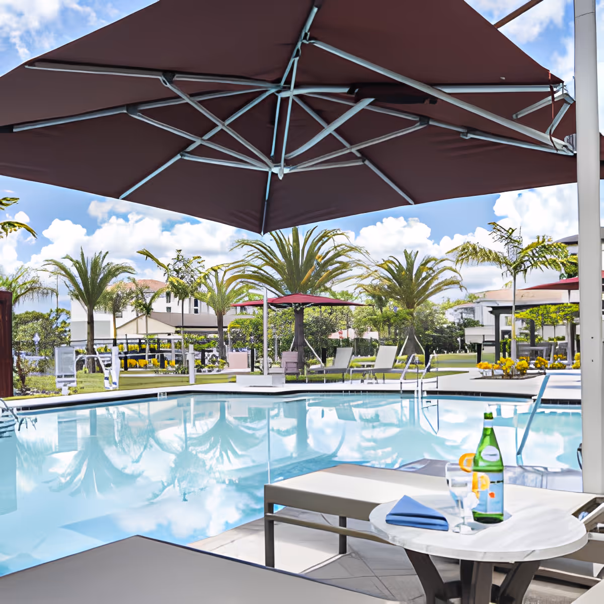 Outdoor swimming pool area with lounge chairs and tables under large umbrellas. Palm trees and greenery surround the pool, with a clear blue sky and some clouds in the background. A bottle of sparkling water and a glass with a lemon slice are on a small round table near the pool.