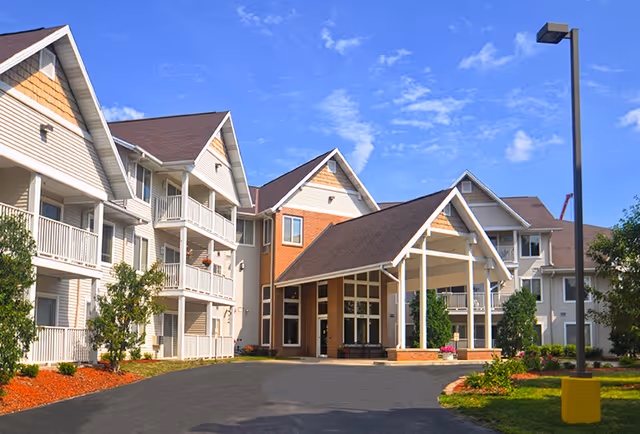 Exterior view of Meadowmere & Mitchell Manor West Allis senior living facility showing a multi-story building with balconies, a covered entrance, landscaped grounds, and a clear blue sky.