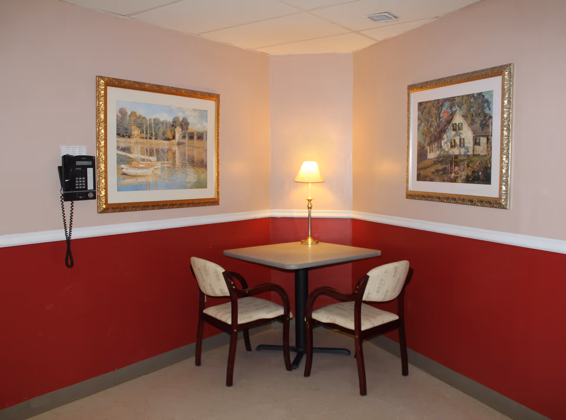 A small corner seating area with a square table and two cushioned chairs. A table lamp with a beige shade is placed on the table, casting a warm light. The walls are painted with a two-tone scheme: the lower half is dark red and the upper half is light beige. Two framed paintings hang on the walls, one depicting a riverside scene with boats and the other showing a house surrounded by trees. A black wall-mounted telephone is visible on the left wall.
