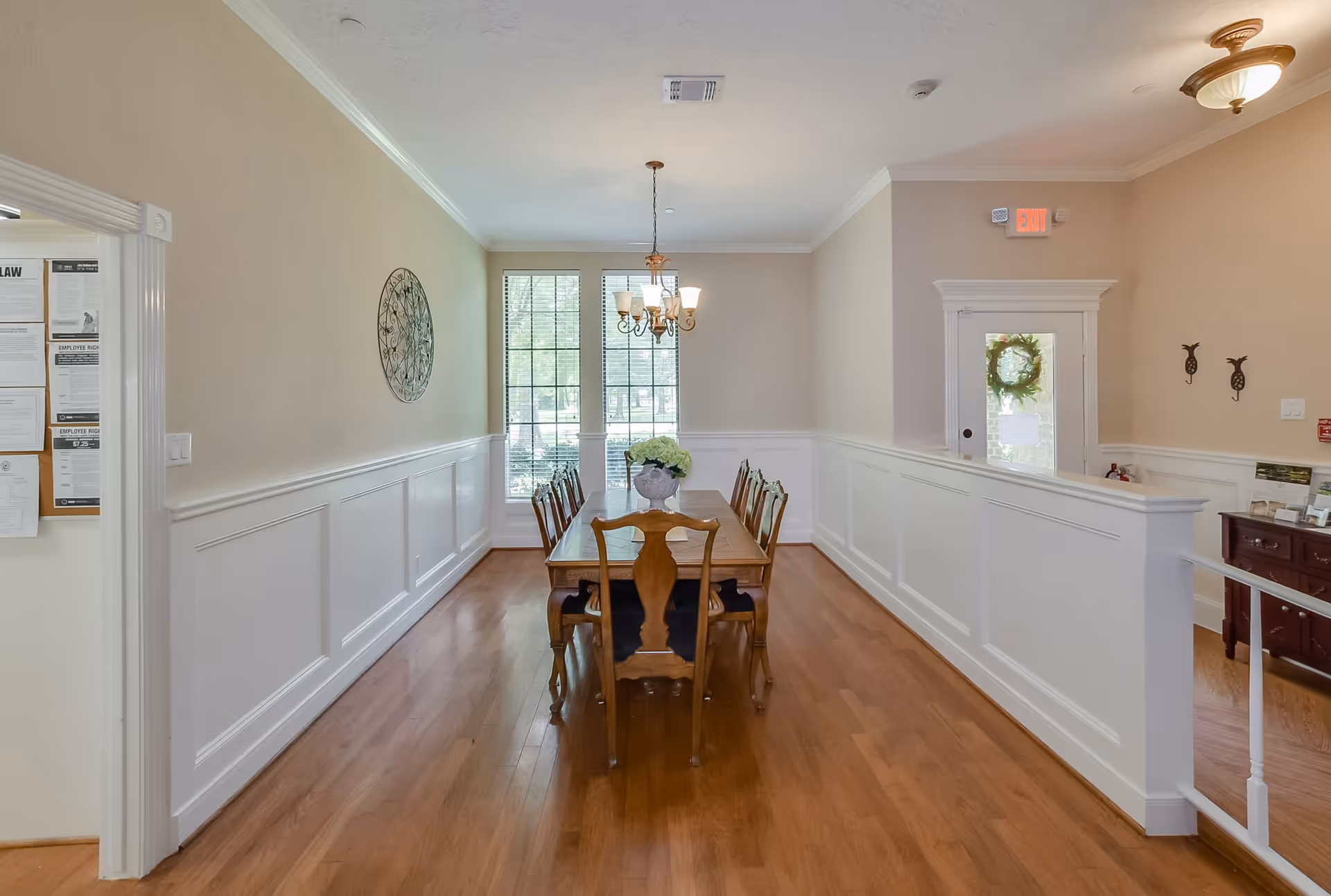 A dining room with a wooden dining table and eight matching chairs. The room has light beige walls with white wainscoting, a chandelier hanging above the table, and two tall windows letting in natural light. There is a decorative metal wall art on the left wall and an exit door with a wreath on it at the far end of the room.