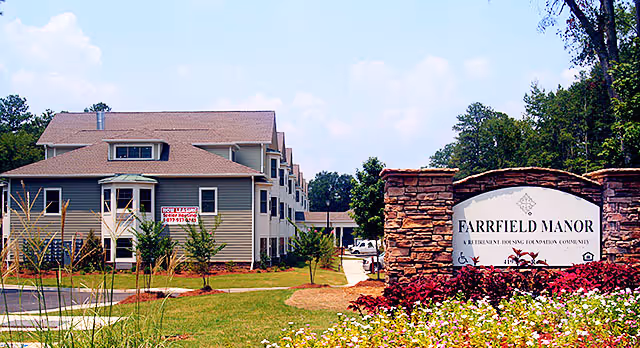 Exterior view of Farrfield Manor Senior Apartments showing a multi-story residential building with beige siding and a brown roof. There is a landscaped lawn with young trees and a stone sign in the foreground that reads 'Farrfield Manor A Retirement Housing Foundation Community.'