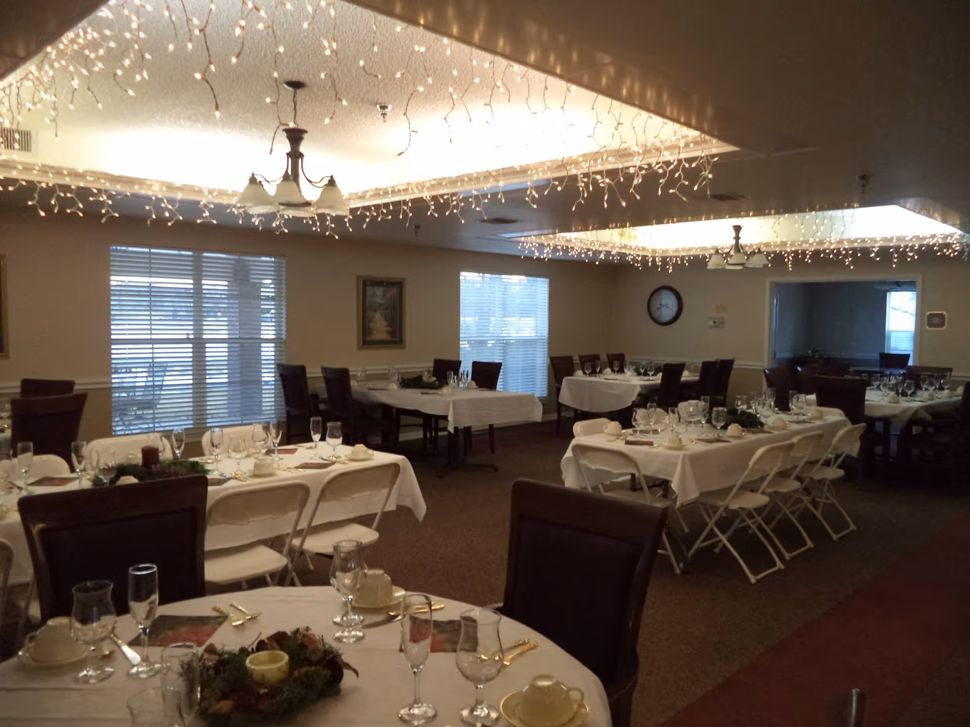 Dining room with multiple tables set for a meal, white tablecloths, chairs and overhead string lights.