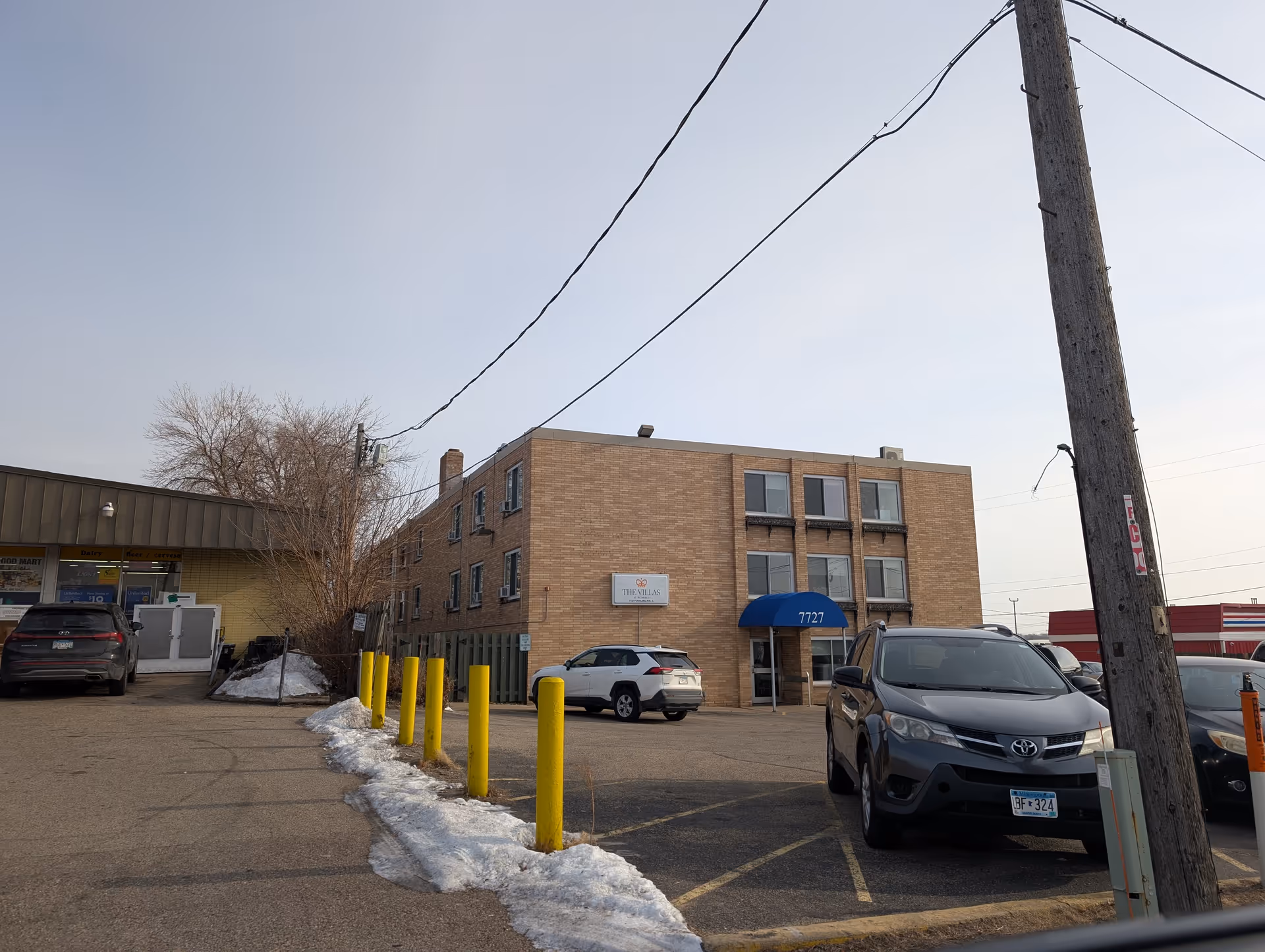 Three-story brick building with a blue awning labeled 7727, parked cars in the front lot and yellow bollards along a snow-dusted driveway.