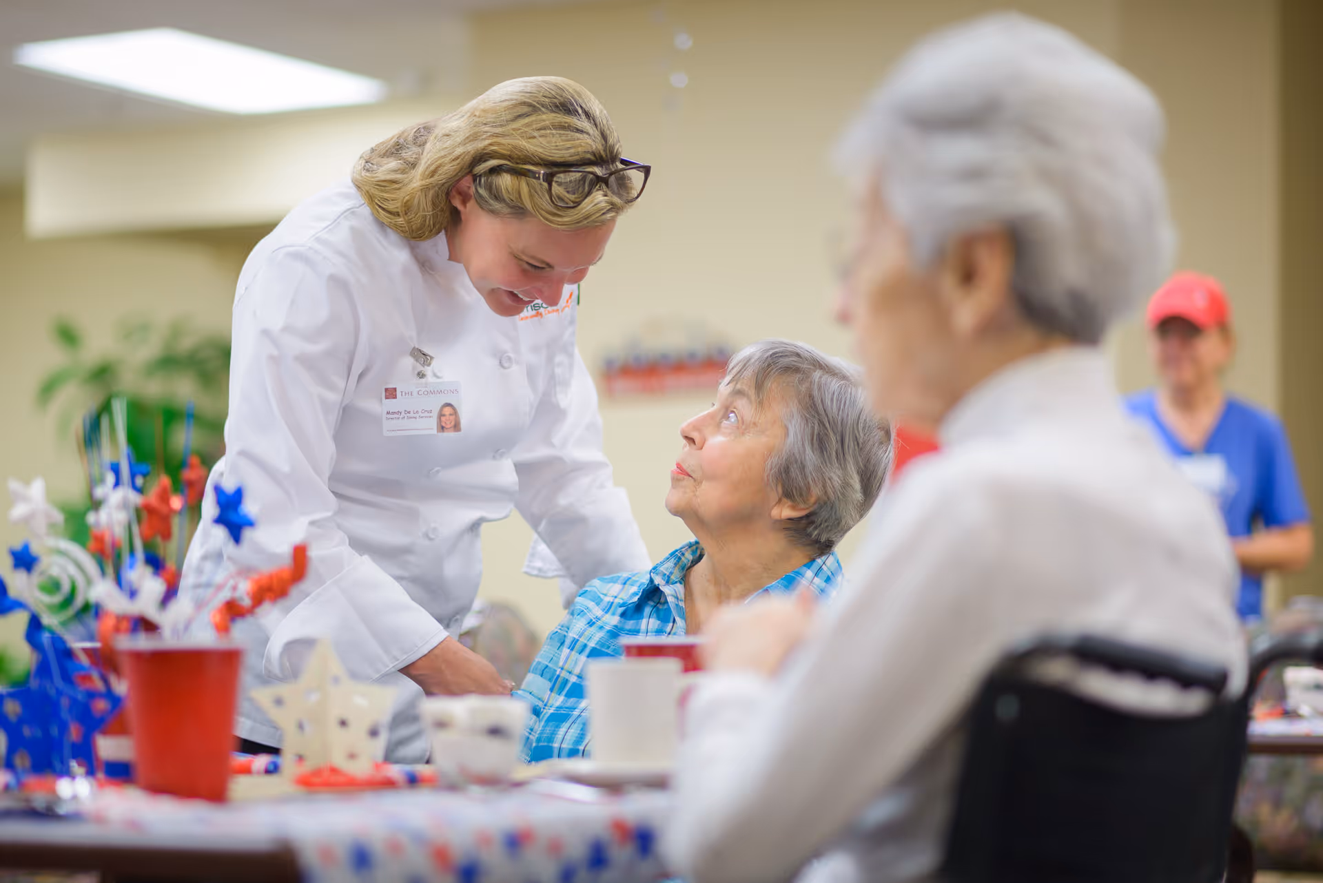 A staff member in a white uniform leans down to speak with a seated elderly woman at a decorated dining table while other residents sit nearby.