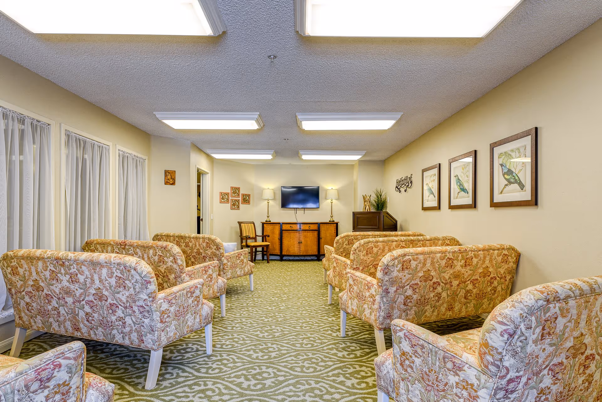 A senior living facility common area with floral patterned armchairs arranged in two rows facing a flat-screen TV mounted on a wooden cabinet. The room has beige walls adorned with framed bird artwork and decorative wall hangings. The floor is covered with a green patterned carpet, and the ceiling has multiple rectangular fluorescent light fixtures. There are windows with sheer white curtains on the left side and a wooden chair and lamps near the TV.
