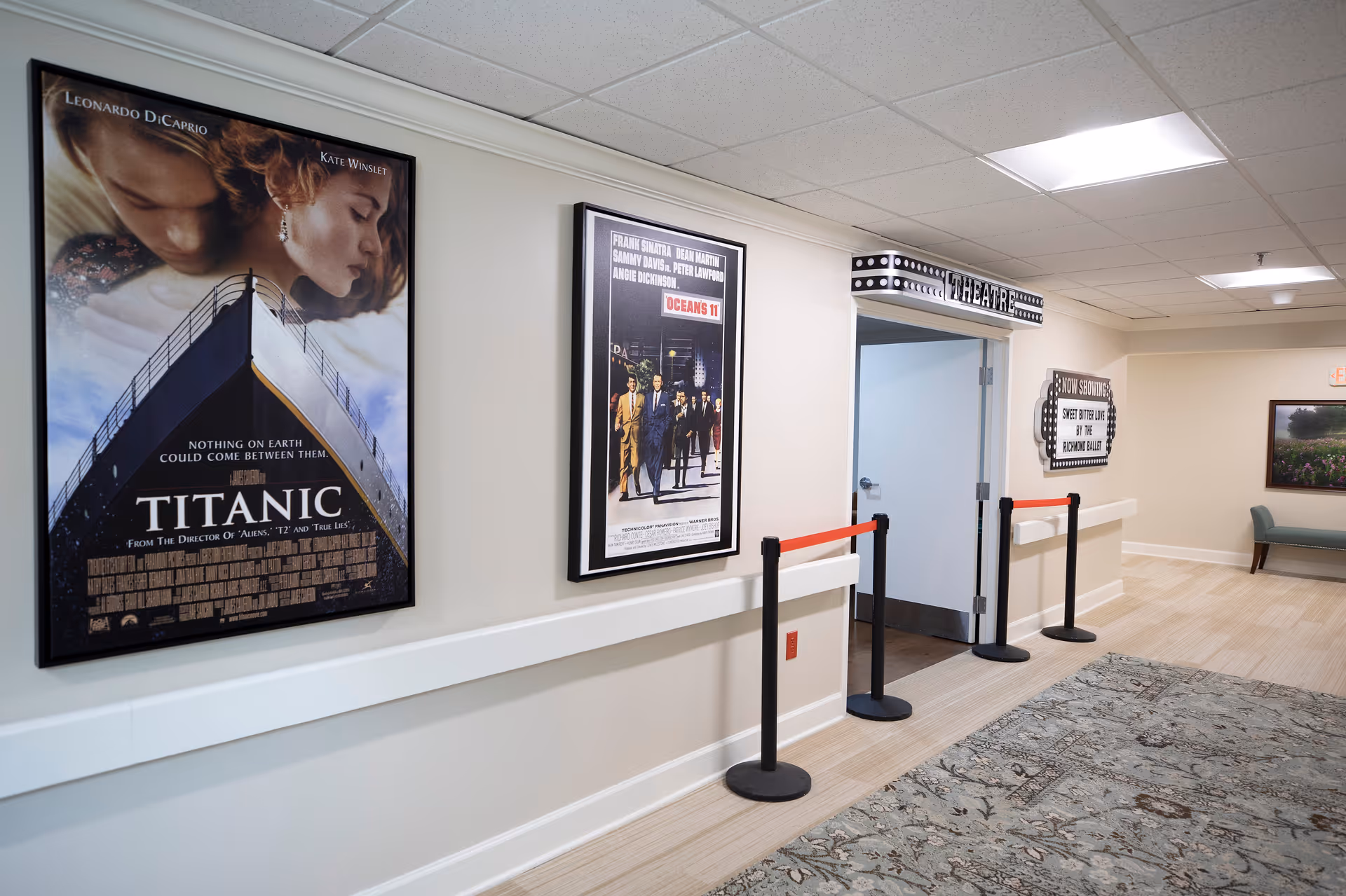 Interior hallway of a retirement community with movie posters for Titanic and Oceans 11 on the wall. There is a theater entrance with a marquee sign above the door and a sign indicating the current movie showing. The hallway has a patterned carpet, light-colored walls, and ceiling lights.