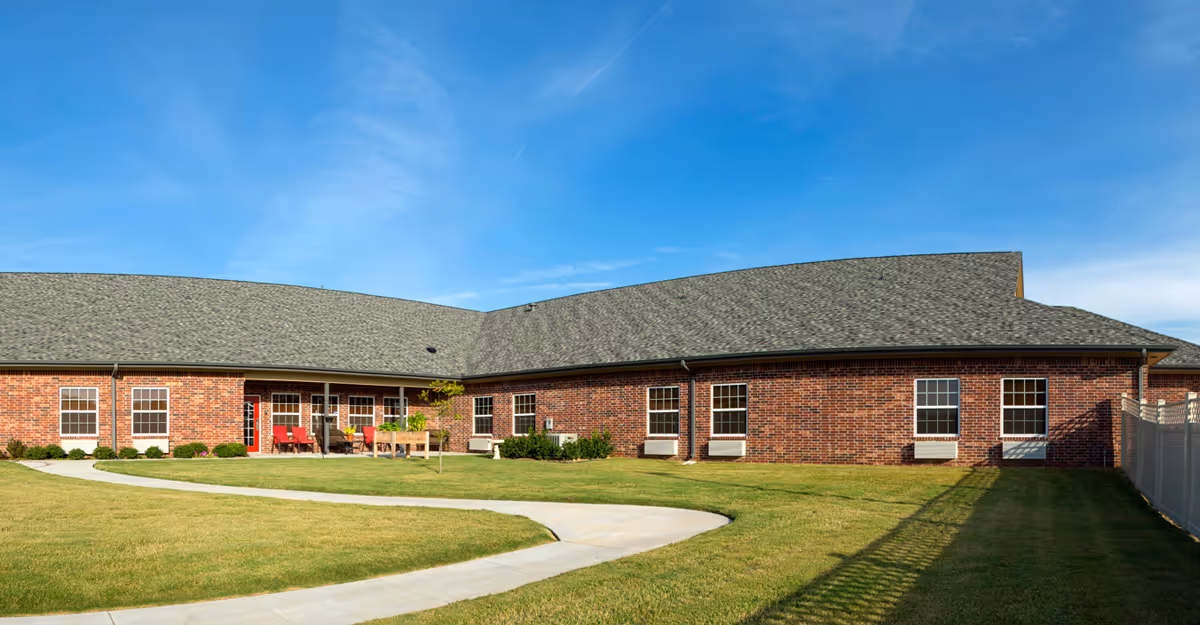Exterior view of a single-story brick building with multiple windows and a gray shingled roof under a clear blue sky. There is a curved concrete pathway leading through a well-maintained grassy lawn in front of the building. Red chairs and a table are visible on a small patio area near the entrance.