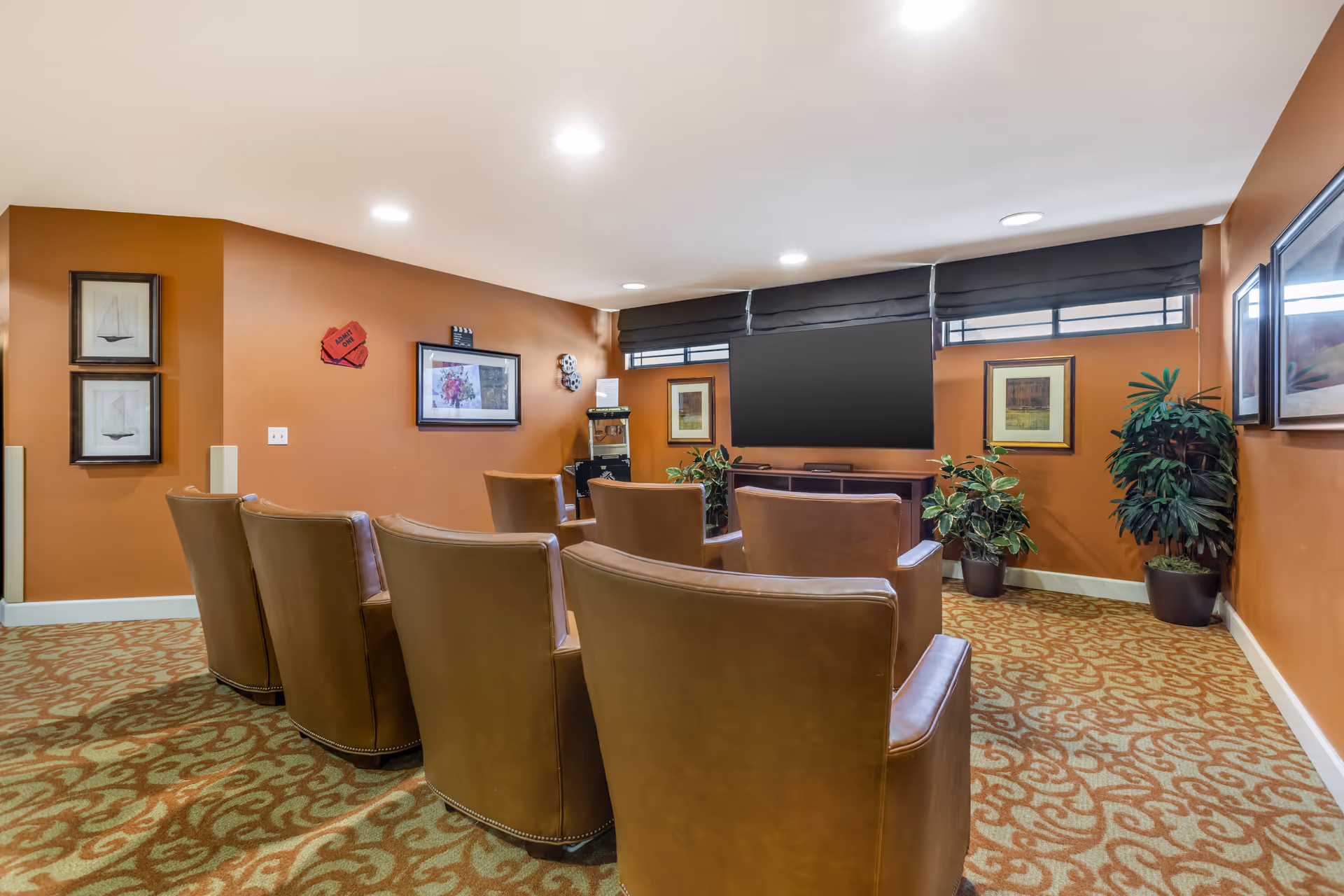 A cozy media room with six brown leather armchairs arranged in two rows facing a large flat-screen TV mounted on an orange wall. The room has patterned carpet flooring, framed artwork on the walls, two potted plants, and a popcorn machine in the corner. The windows have black roller shades partially drawn.