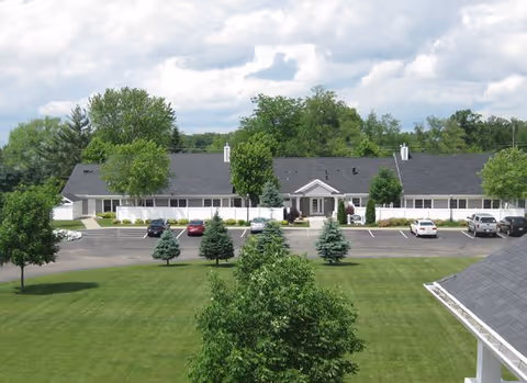 Exterior view of a single-story senior living facility building with a dark roof and white walls, surrounded by green trees and a well-maintained lawn. Several cars are parked in the parking lot in front of the building under a partly cloudy sky.