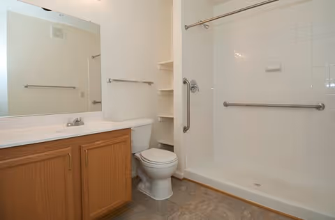 A clean bathroom featuring a white toilet, a wooden vanity with a white countertop and sink, a large mirror above the sink, and a white bathtub with grab bars and a shower curtain rod.