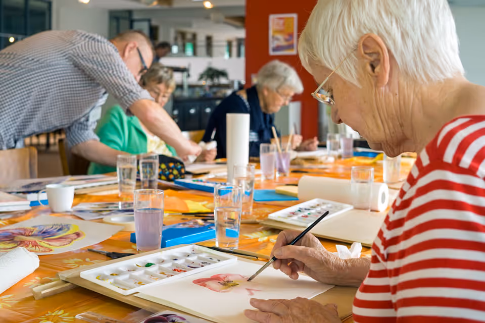 A group of elderly people engaged in a watercolor painting activity around a table covered with an orange floral tablecloth. One woman in the foreground wearing glasses and a red and white striped shirt is painting a flower on paper. Various art supplies, water glasses, and paper towels are scattered on the table.