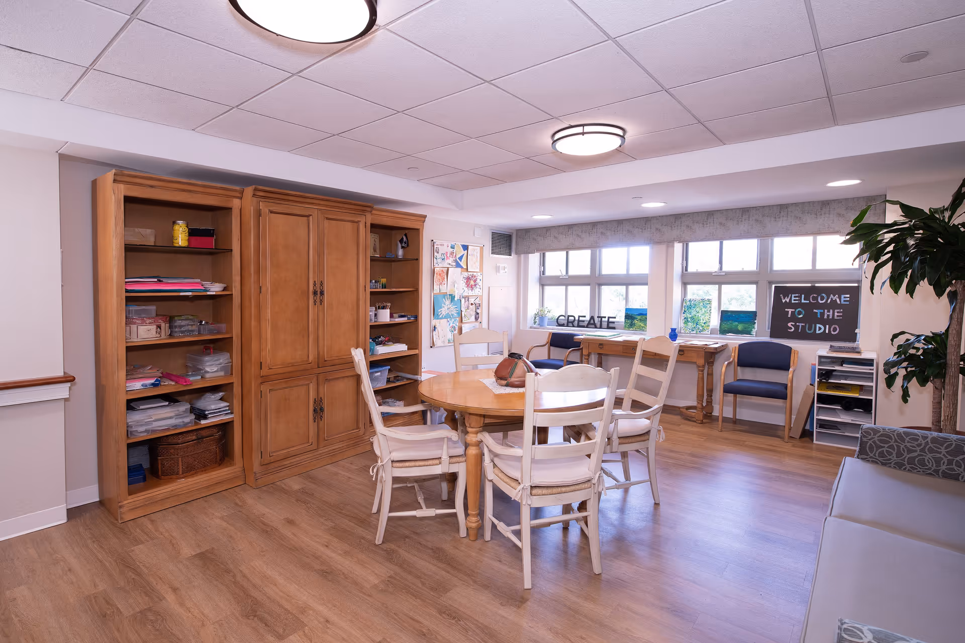 A bright and spacious activity room with wooden flooring, a round wooden table surrounded by four white cushioned chairs, wooden shelves filled with craft supplies, and a bulletin board displaying artwork. Large windows let in natural light, and a sign on the wall reads 'WELCOME TO THE STUDIO' with another sign on the windowsill saying 'CREATE'. There are additional chairs and a potted plant in the room.