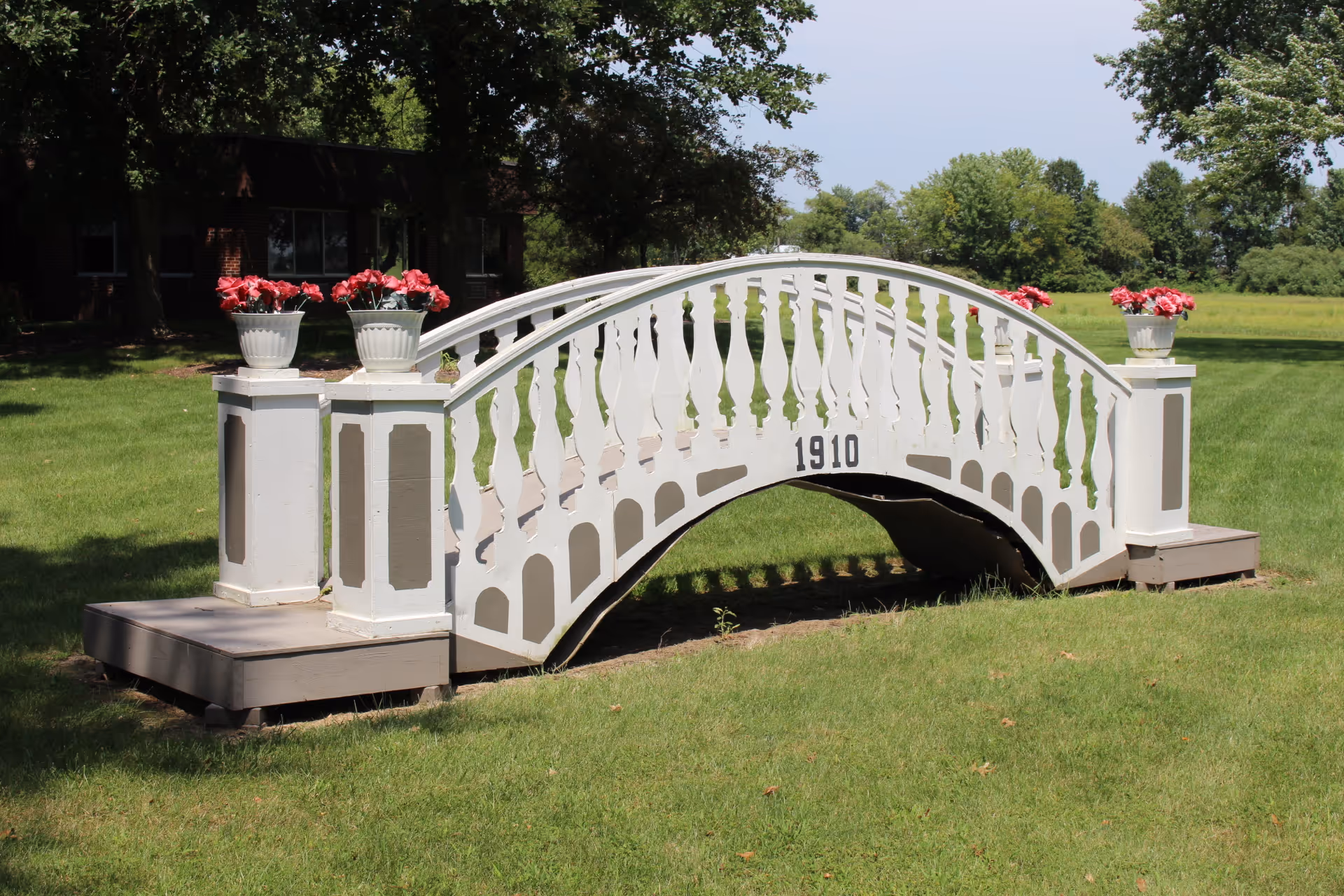 A small decorative white wooden bridge with the number 1910 on its side, placed on a grassy lawn with flower pots containing red flowers on each corner. Trees and a building are visible in the background under a clear sky.
