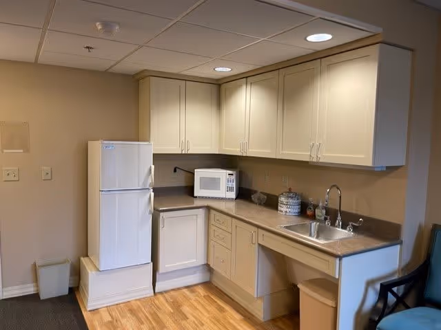A small kitchen area with beige cabinets, a white refrigerator on a raised platform, a microwave on the countertop, a stainless steel sink with a faucet, and a trash bin underneath the counter. The floor is wooden, and there is a blue chair partially visible on the right side.