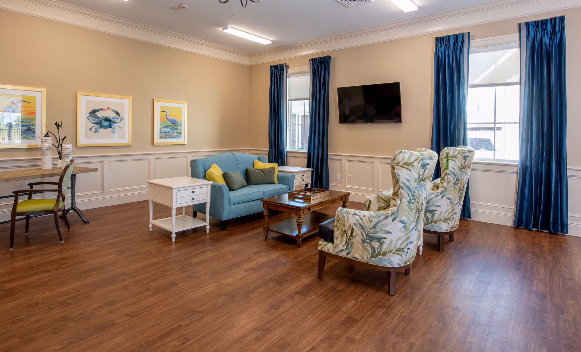 Bright communal living room with a blue sofa, patterned armchairs, coffee table, and wall-mounted TV.