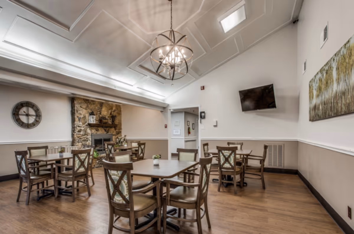 A bright communal dining room with wooden tables and chairs, a stone fireplace, chandelier, and wall-mounted TV.