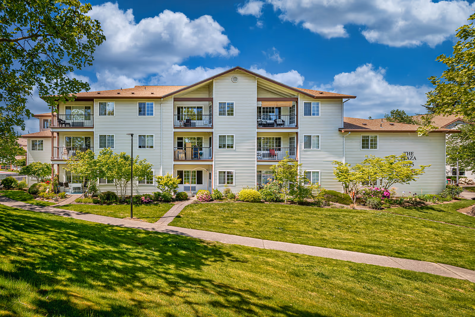 Exterior view of a three-story residential building with balconies, surrounded by well-maintained green lawns, trees, and shrubs under a partly cloudy blue sky.