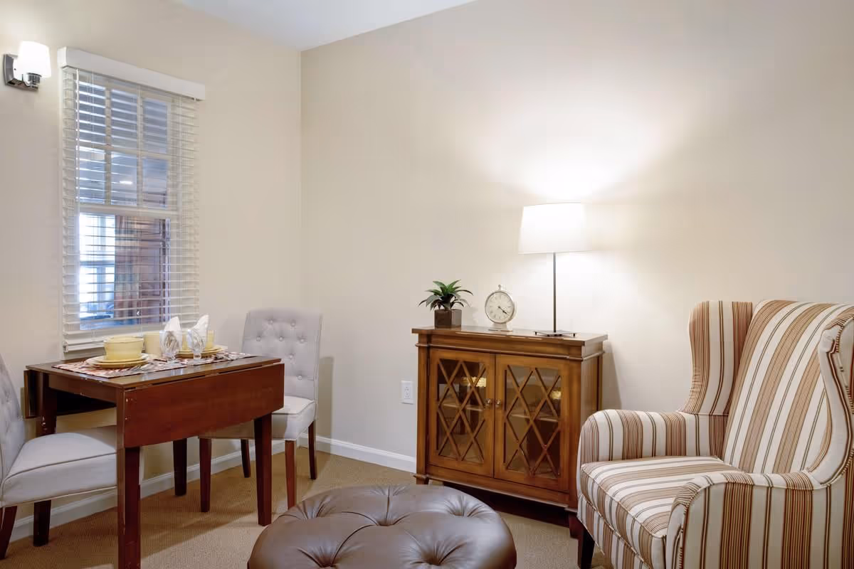 A cozy interior room with a small wooden dining table set for two with plates, cups, and napkins. Two upholstered chairs are placed at the table. Next to the table is a wooden cabinet with glass doors, topped with a small plant, a clock, and a lit table lamp. A striped armchair and a round leather ottoman are also visible. The room has beige walls and a window with white blinds.