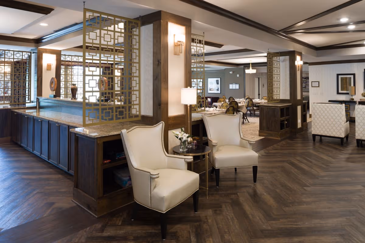 A spacious and elegant interior common area with two cream-colored armchairs and a small round table with flowers between them. The room features dark wood flooring, decorative wooden dividers with geometric patterns, and warm lighting. In the background, there are dining tables and chairs arranged neatly, suggesting a dining or lounge area within a senior living facility.