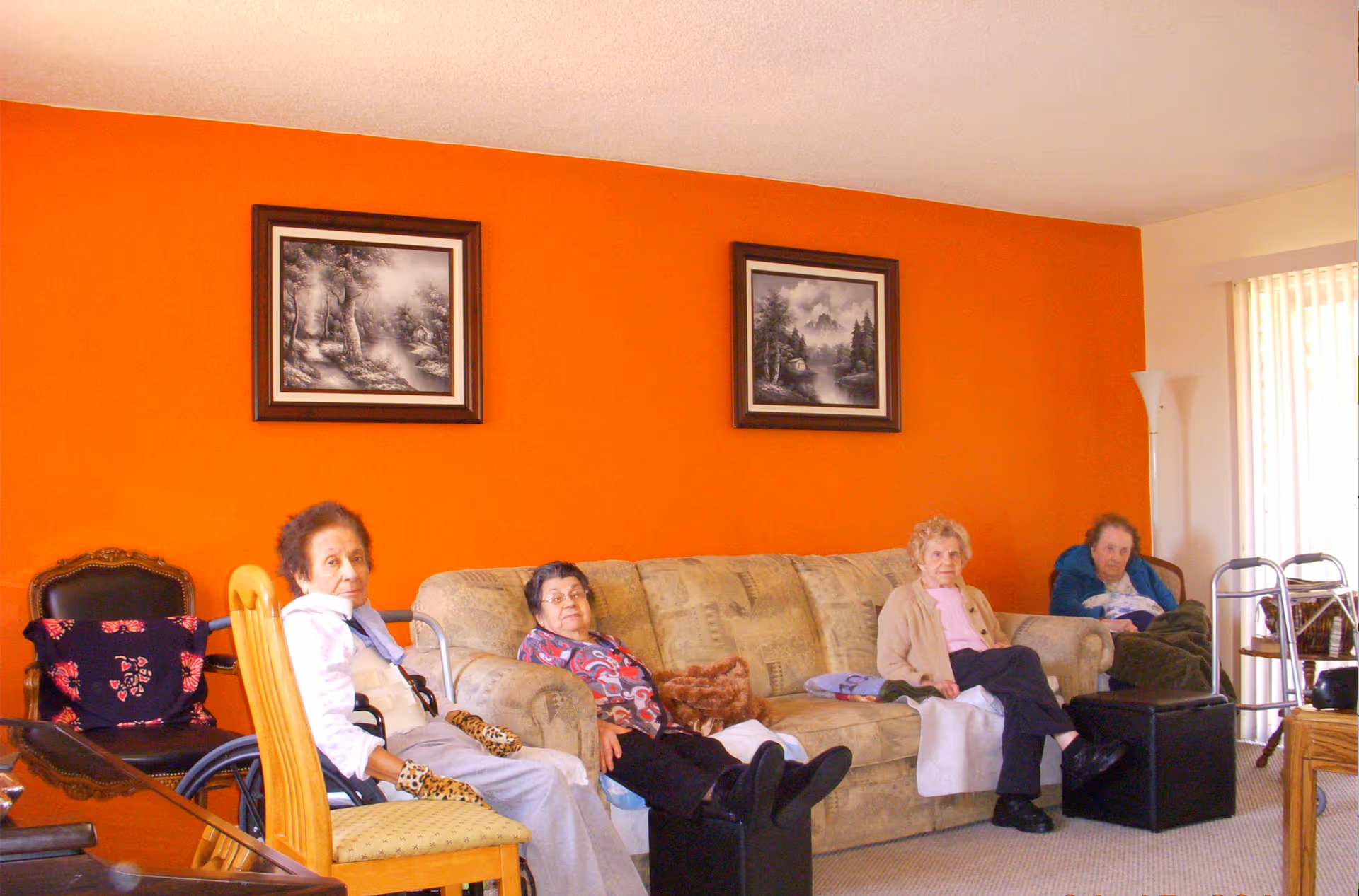 Four elderly women sitting in a living room with an orange accent wall behind them. Two framed black and white landscape pictures hang on the wall. The women are seated on a beige couch and chairs, with some mobility aids like a walker visible nearby. The room has a carpeted floor and vertical blinds covering a window or sliding door.