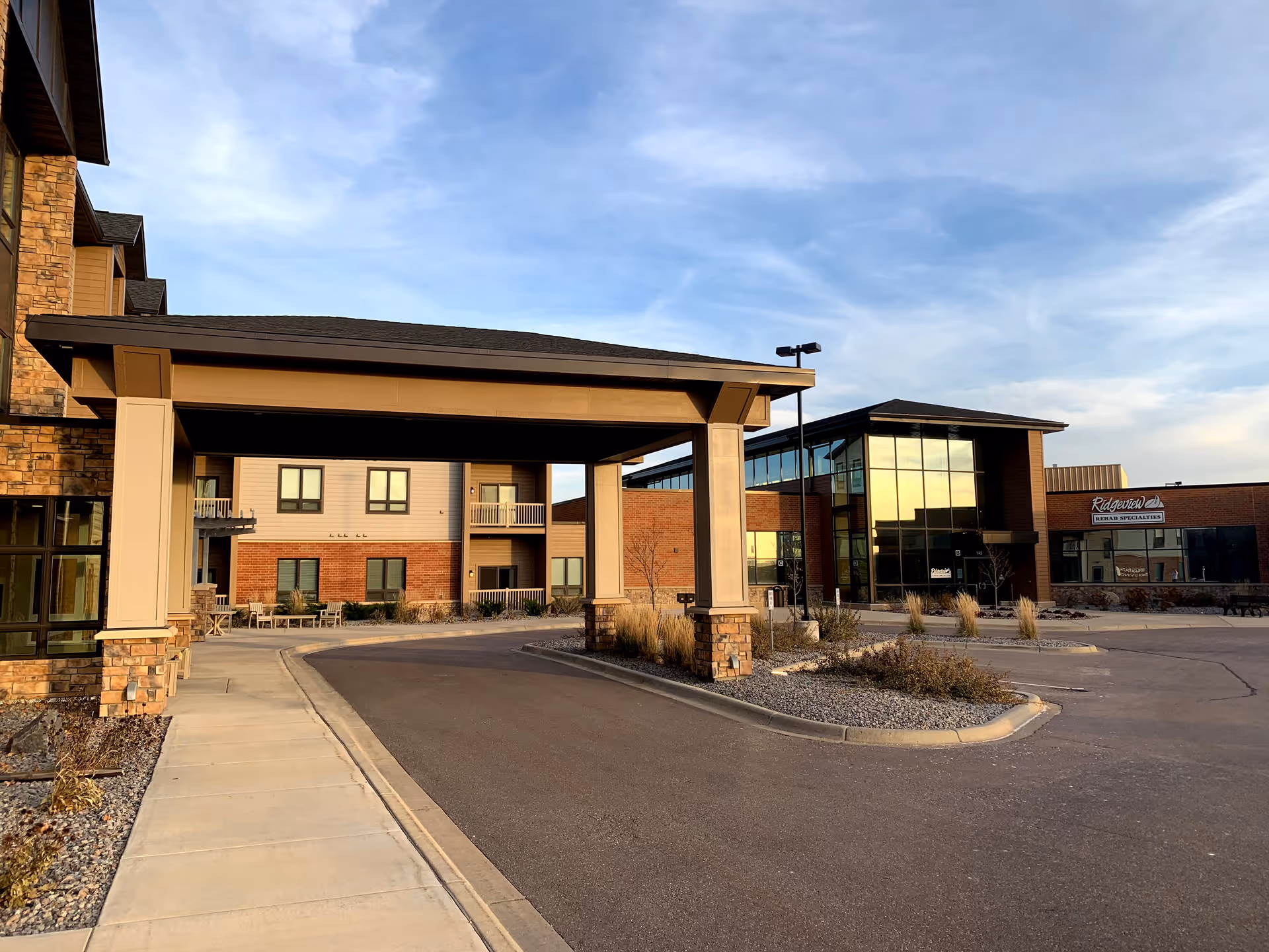 Exterior view of a senior living facility with a covered driveway entrance, multi-story building with brick and beige siding, large windows, and a clear sky in the background.
