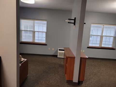 An interior room with two large windows with blinds, light blue walls, and brown carpet. There is a wooden cabinet against a partial wall in the center, and a small heater or air conditioning unit beneath one window. The room appears empty and well-lit with ceiling lights.