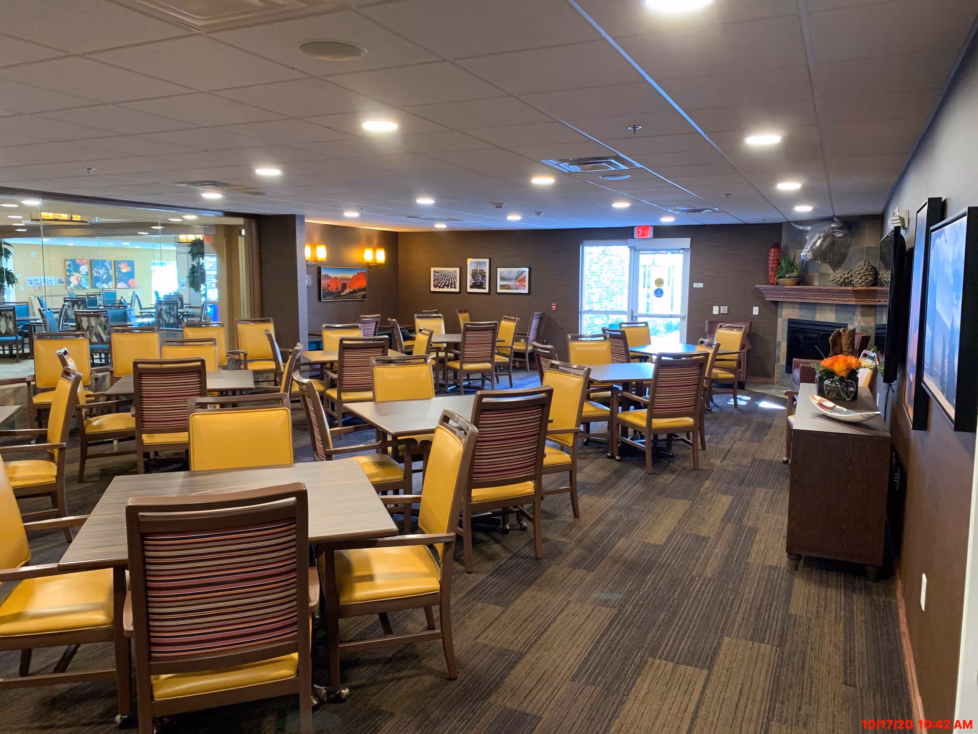 Dining room with multiple tables and yellow-cushioned chairs, a fireplace, and wall art in a senior living facility.